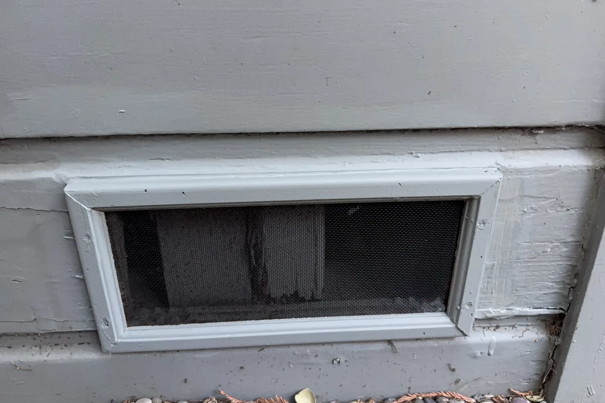 Foundation vent on gray wood siding retrofitted with ember-resistant fine mesh inside a painted trim frame