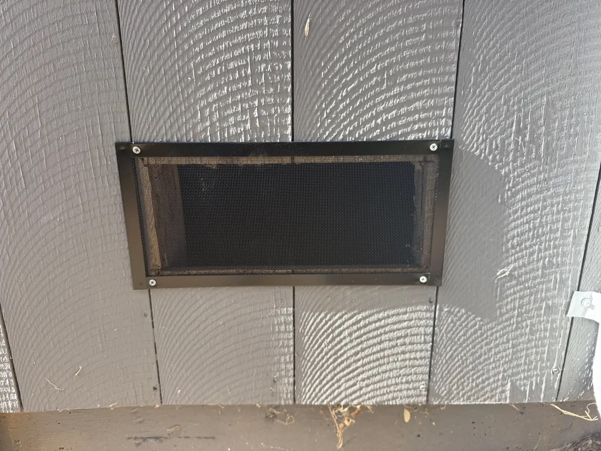 Foundation vent on gray wood siding fitted with a new ember-resistant mesh vent screen