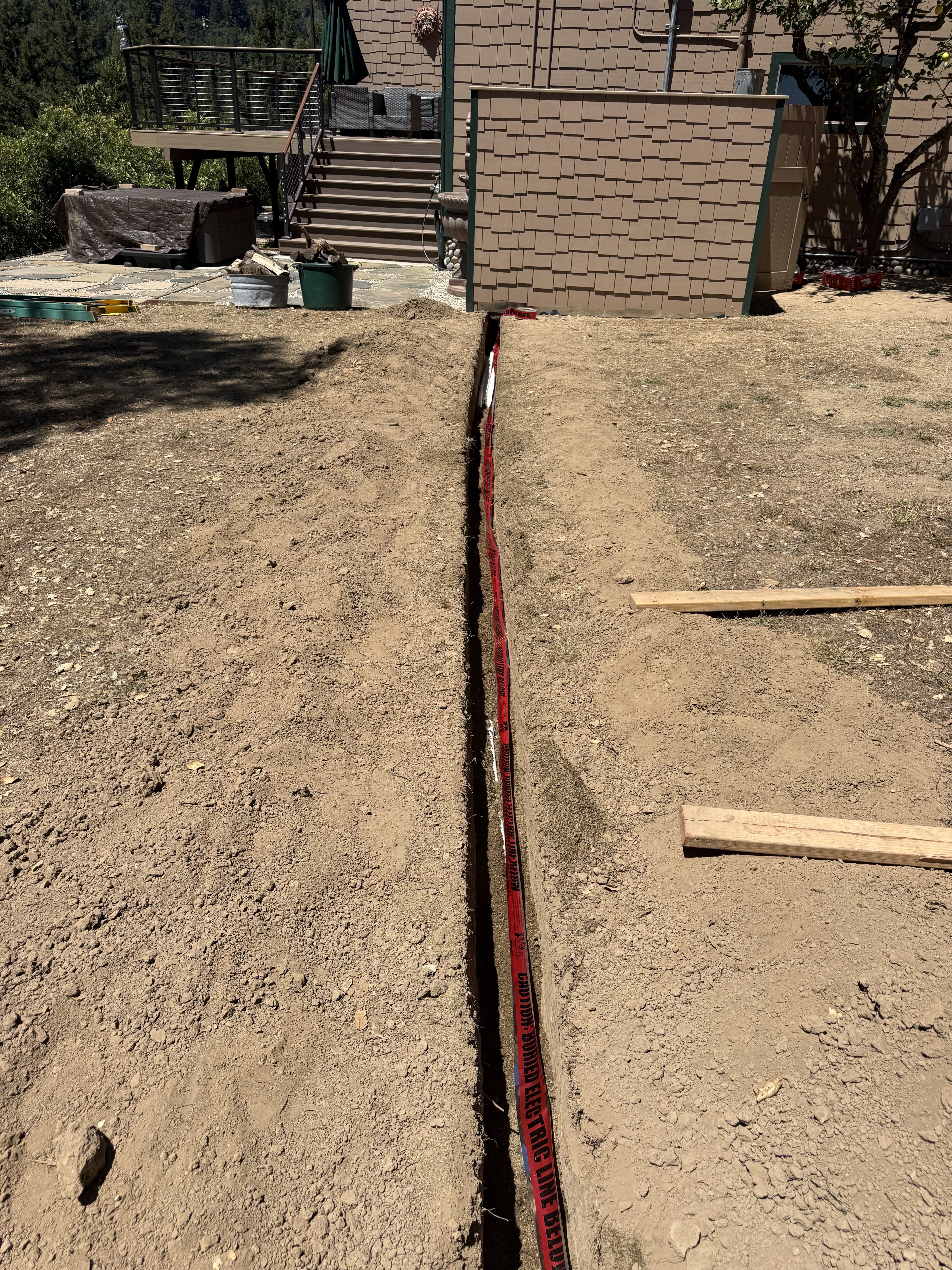 Trenched water supply line with caution tape running across the yard to the Summit Road home, connecting to new 10,000-gallon water storage tanks