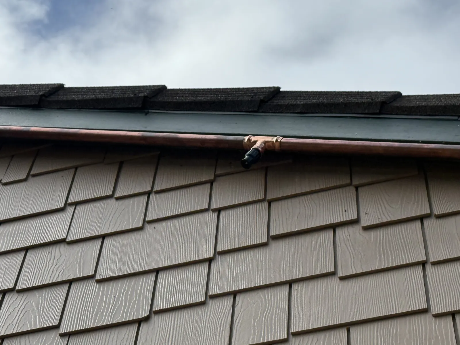Close-up of a FireRoofs copper supply line with a sprinkler nozzle on a tee fitting mounted cleanly along the roofline above shake siding