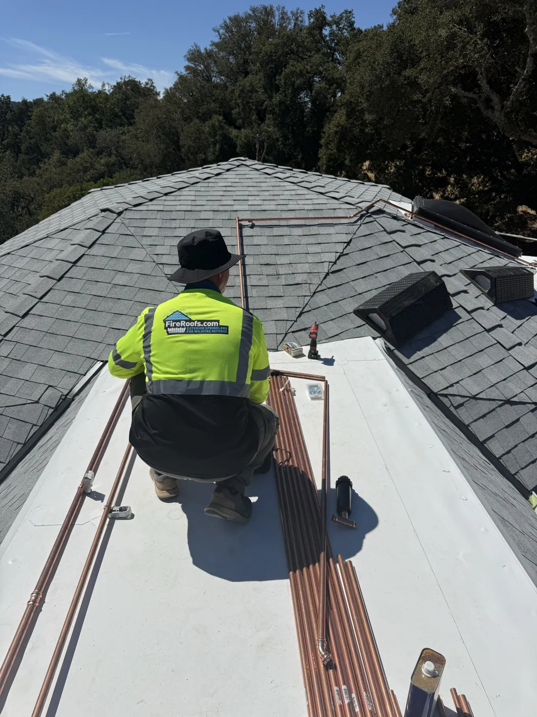 FireRoofs Vetted Installer staging copper sprinkler lines on a shingle roof surrounded by oaks