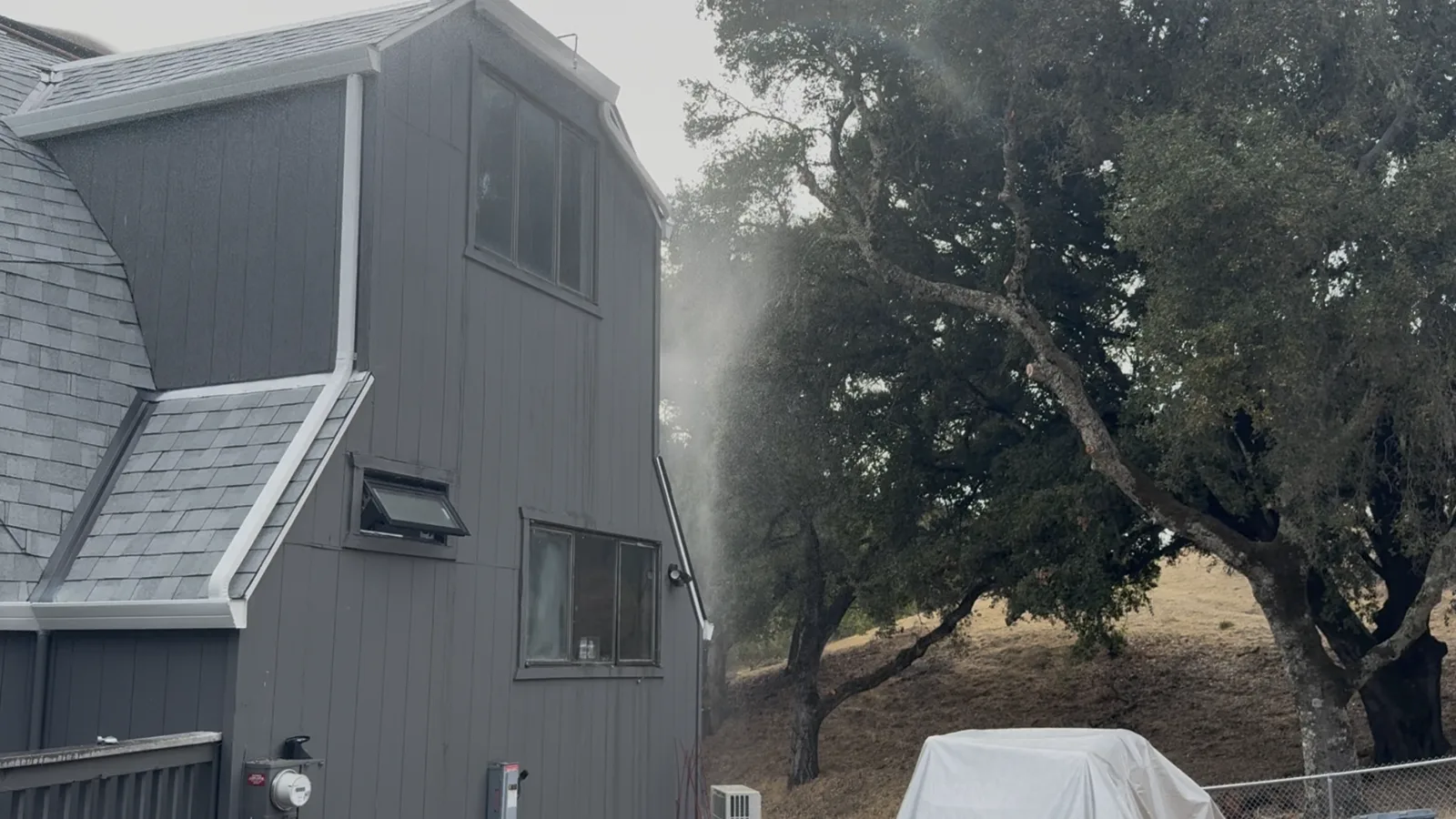 Exterior sprinkler spraying toward oak trees and dry grass beside a home