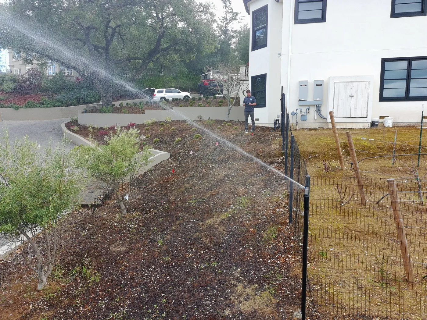 Perimeter sprinkler on a steel post spraying across a landscaped hillside beside a home