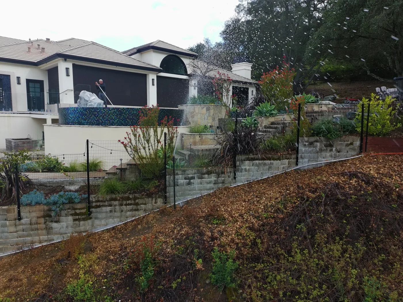 Perimeter sprinklers on steel posts wetting a terraced hillside garden below a home
