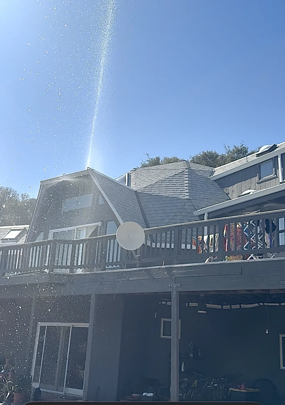 Roof sprinkler throwing a high arc of water above a hillside home with a large deck